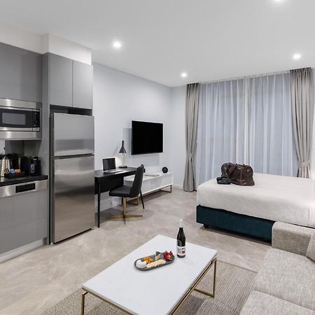 Meriton Suites Sussex Street, Sídney Exterior foto The photo shows a modern studio apartment. On one side, there is a kitchenette with sleek gray cabinets, a refrigerator, and built-in appliances. In the living area, there is a comfortable gray sofa, a white coffee table with a fruit platter and a bo