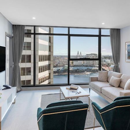 Meriton Suites Sussex Street, Sídney Exterior foto The photo shows a modern living room with large windows that offer a view of the cityscape and water. The interior features a light color palette with pale blue walls and soft lighting. There is a comfortable gray sofa and two green accent chairs, ar