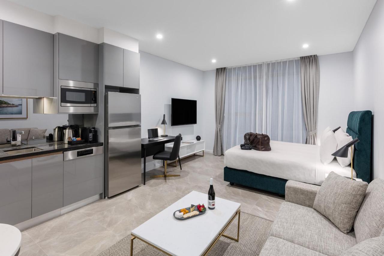 Meriton Suites Sussex Street, Sídney Exterior foto The photo shows a modern studio apartment. On one side, there is a kitchenette with sleek gray cabinets, a refrigerator, and built-in appliances. In the living area, there is a comfortable gray sofa, a white coffee table with a fruit platter and a bo