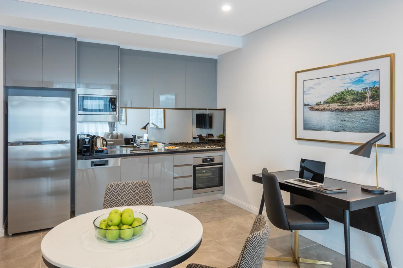 Meriton Suites Sussex Street, Sídney Exterior foto The photo shows a modern kitchen and dining area in an apartment. The kitchen features sleek gray cabinetry, a stainless steel fridge, and a microwave. There is a countertop with cooking appliances, including an oven and stovetop. A small round dinin