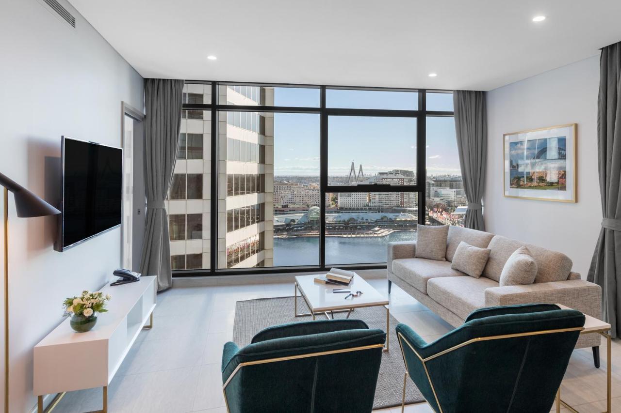 Meriton Suites Sussex Street, Sídney Exterior foto The photo shows a modern living room with large windows that offer a view of the cityscape and water. The interior features a light color palette with pale blue walls and soft lighting. There is a comfortable gray sofa and two green accent chairs, ar