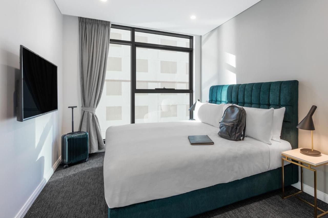 Meriton Suites Sussex Street, Sídney Exterior foto The photo shows a modern hotel room featuring a comfortable bed with white bedding and a teal-colored headboard. There is a neatly placed black backpack on the bed, along with a small notebook or tablet. A television is mounted on the wall opposite t