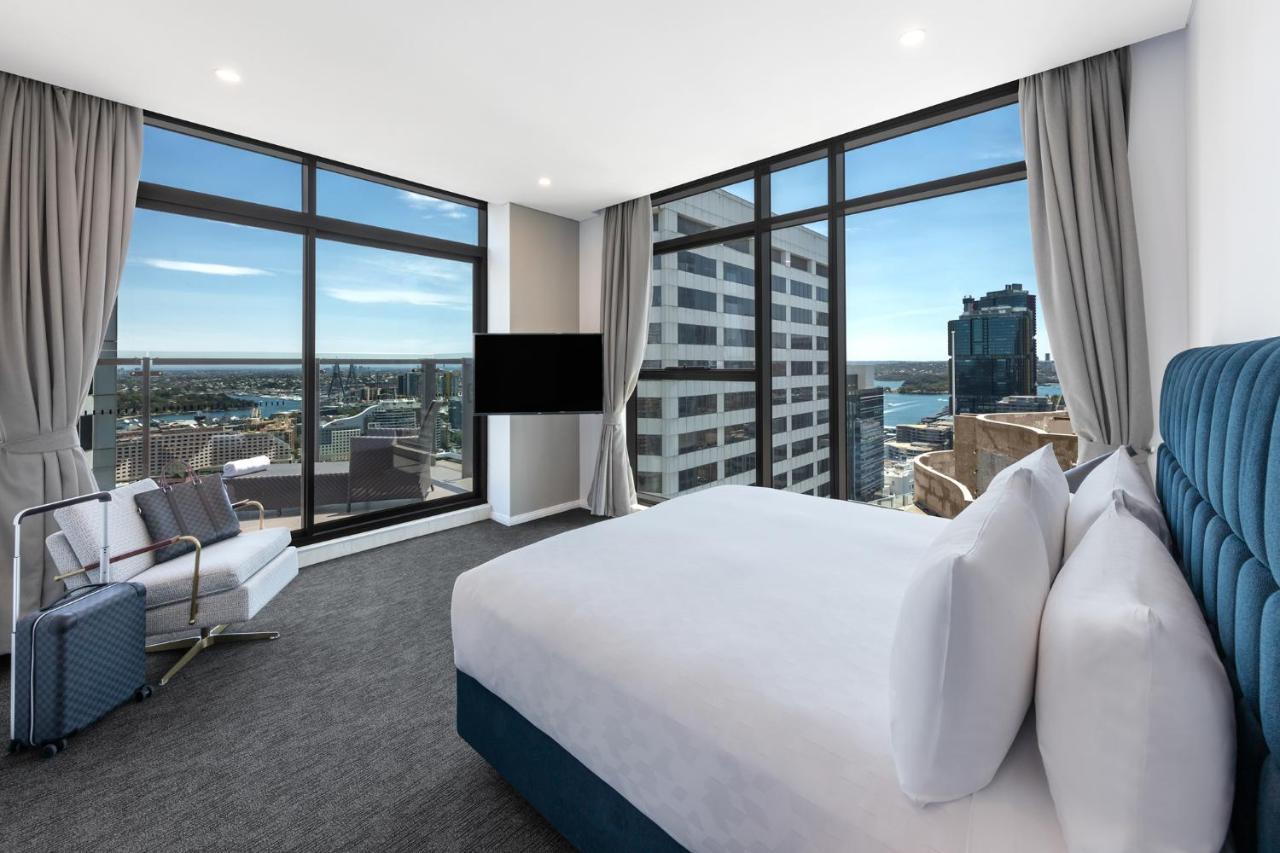 Meriton Suites Sussex Street, Sídney Exterior foto The photo shows a modern hotel room featuring large windows that provide a panoramic view of the cityscape and waterfront. The room is furnished with a king-size bed dressed in white linens, a comfortable armchair, and a flat-screen TV mounted on the