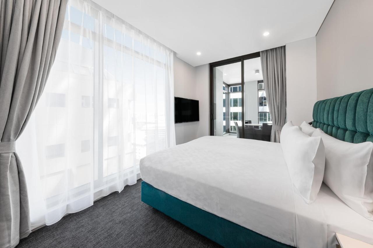 Meriton Suites Sussex Street, Sídney Exterior foto The photo shows a modern bedroom featuring a large bed with a white duvet and pillows. The headboard is turquoise, adding a pop of color to the neutral-toned room. There are sheer white curtains covering large windows that let in natural light, creat