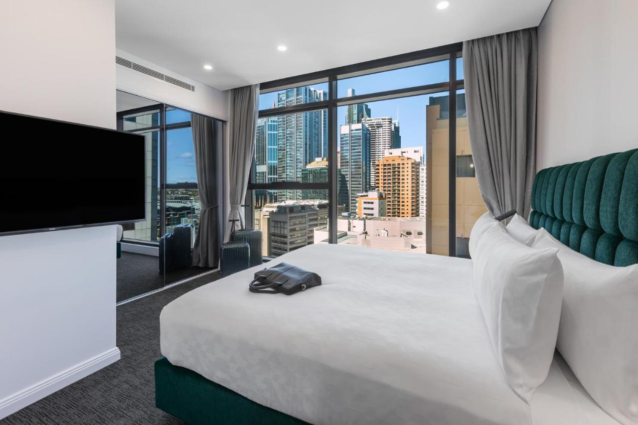 Meriton Suites Sussex Street, Sídney Exterior foto The photo shows a modern hotel room featuring a large bed with white linens and a green upholstered headboard. There is a sleek television mounted on the wall opposite the bed, and the room has large windows that provide a view of a cityscape, includ