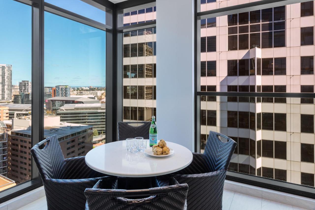 Meriton Suites Sussex Street, Sídney Exterior foto The photo shows a modern dining space with a round white table set for four. There are four black woven chairs around the table. On the table, there's a bottle of sparkling water, two glasses, and a small plate with several cookies. The large windows