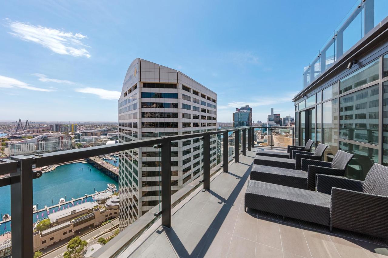 Meriton Suites Sussex Street, Sídney Exterior foto The photo shows a balcony or terrace with a panoramic view of a city skyline. The balcony features modern outdoor furniture, including sleek lounge chairs. In the background, you can see a waterfront area with buildings and a harbor, along with a cle