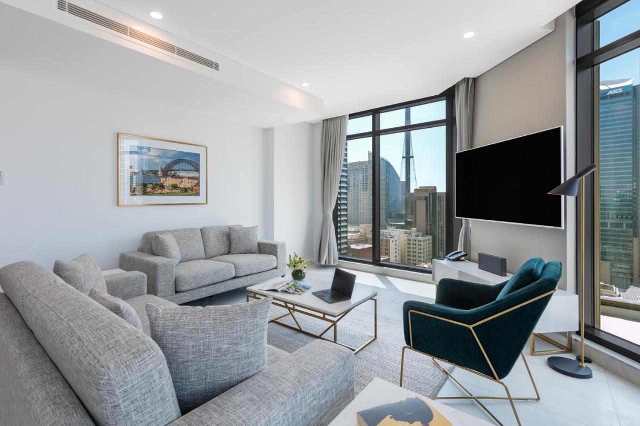 Meriton Suites Sussex Street, Sídney Exterior foto The photo shows a modern living room with large windows offering a view of a city skyline. The room features two gray sofas and a stylish green accent chair. A sleek coffee table is placed in front of the sofas, alongside a laptop. There’s a flat-scr