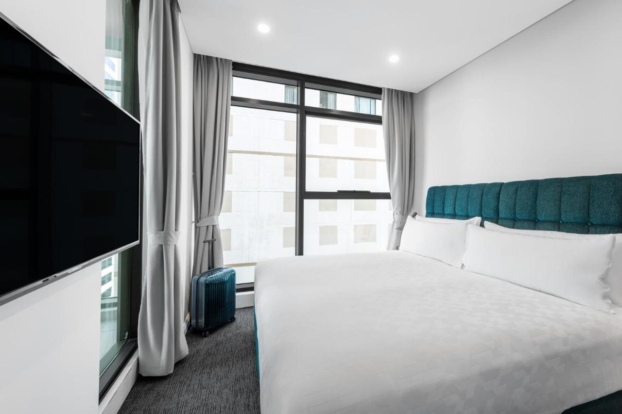 Meriton Suites Sussex Street, Sídney Exterior foto The photo shows a modern hotel room featuring a large bed with a light-colored duvet and a blue upholstered headboard. There are two pillows on the bed. A large window allows natural light to enter the room, and gray curtains frame the window. Next t