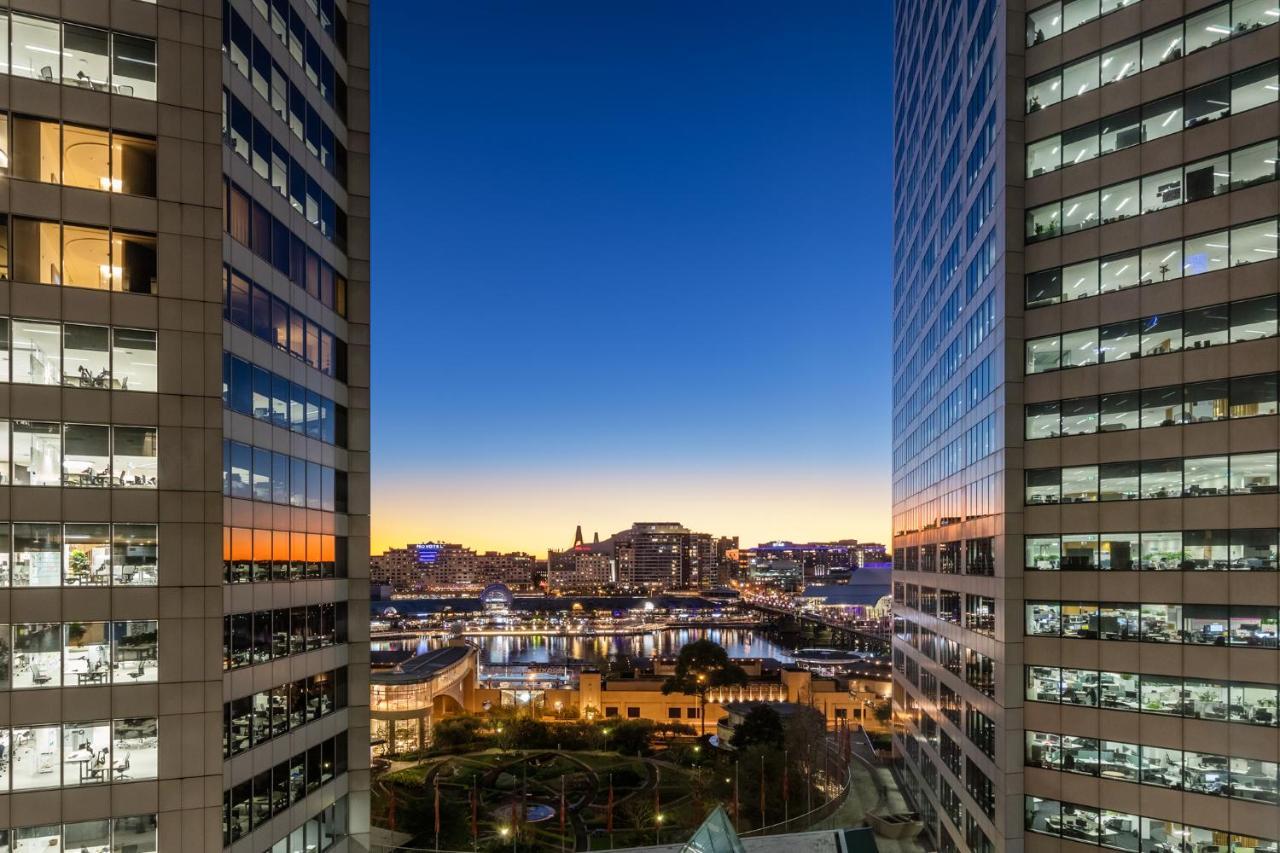 Meriton Suites Sussex Street, Sídney Exterior foto The photo shows a view between two tall buildings, likely office towers, during twilight. On one side, there are glass windows reflecting the evening sky that transitions from blue to orange near the horizon. In the background, a cityscape is visible