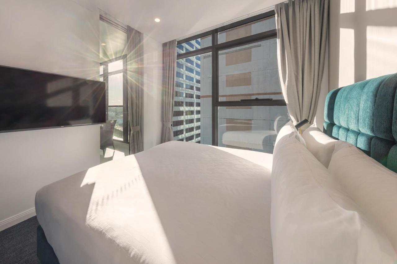 Meriton Suites Sussex Street, Sídney Exterior foto The photo shows a modern hotel room with a neatly made bed featuring white bedding and a plush, tufted headboard in a shade of blue. Natural light is streaming through large windows, which are covered partially by light gray curtains. A flat-screen t