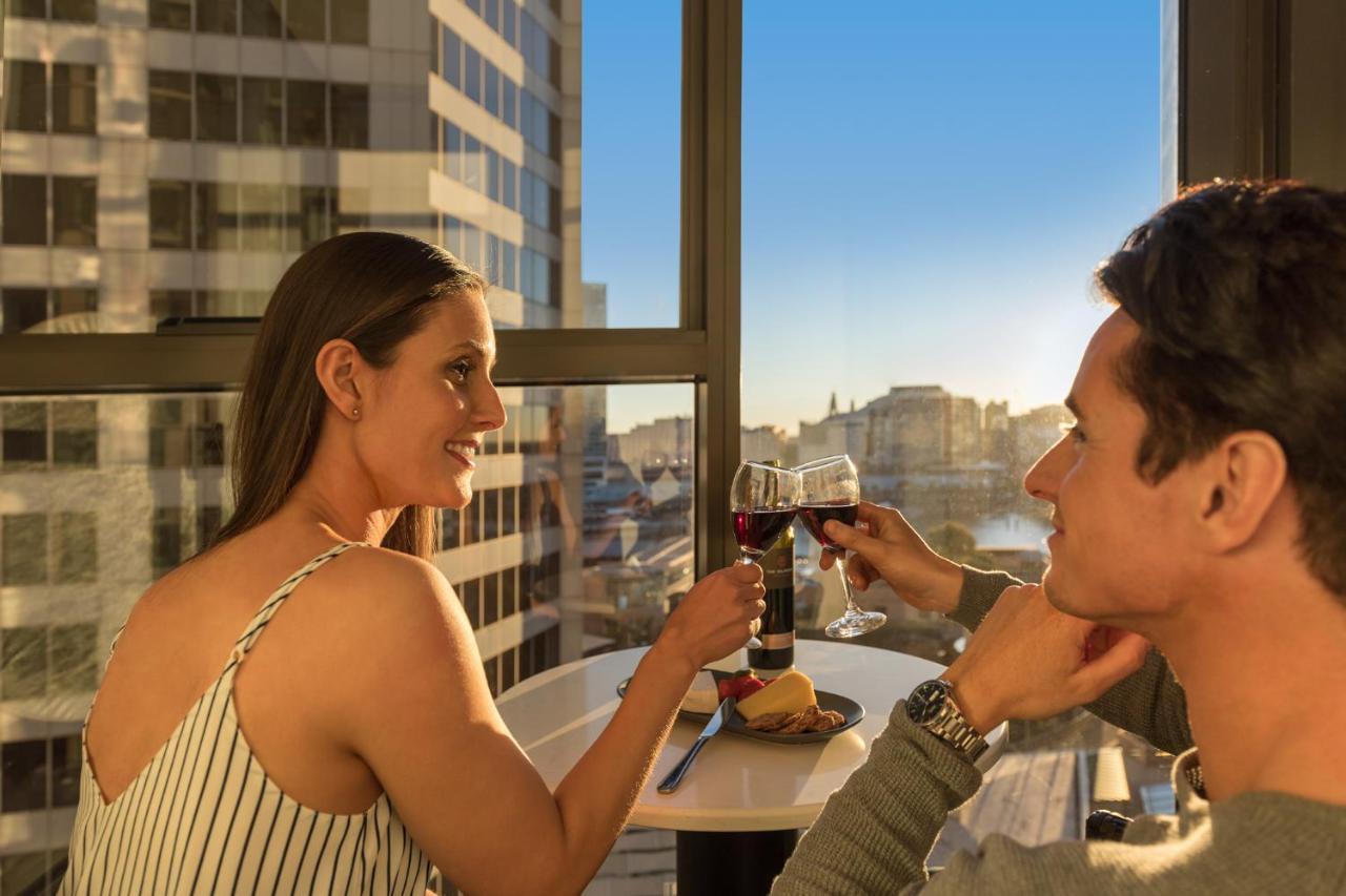 Meriton Suites Sussex Street, Sídney Exterior foto The photo depicts a couple enjoying a drink together in a modern setting. They are sitting at a small table near a large window, which provides a view of a cityscape and a sunset. Both individuals are smiling and engaged in a cheerful toast with glas