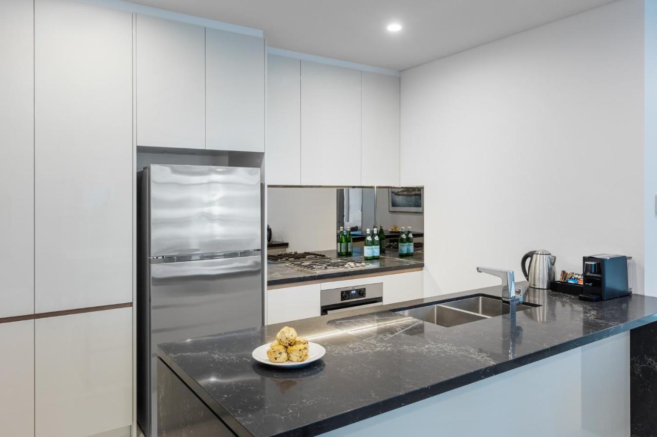 Meriton Suites Sussex Street, Sídney Exterior foto The photo shows a modern kitchen with a sleek, minimalist design. Key features include:

- A shiny stainless steel refrigerator on the left.
- A mirrored backsplash above the stove, which has a gas burner.
- A dark countertop made of what appears to 