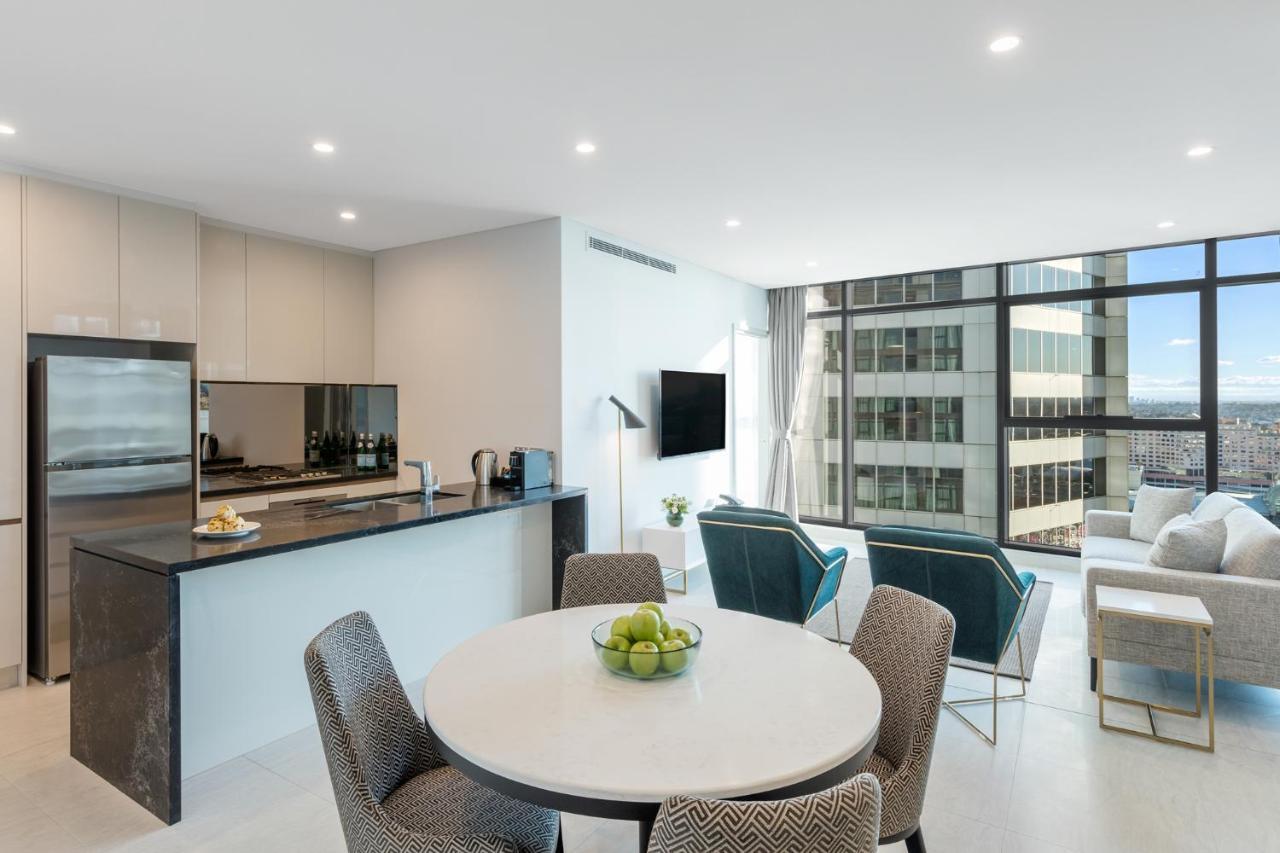 Meriton Suites Sussex Street, Sídney Exterior foto The photo shows a modern and stylish interior space, likely a living room and kitchen area. The kitchen features sleek cabinetry and a countertop, with appliances visible, including a coffee maker. There is a round dining table with gray chairs, set 