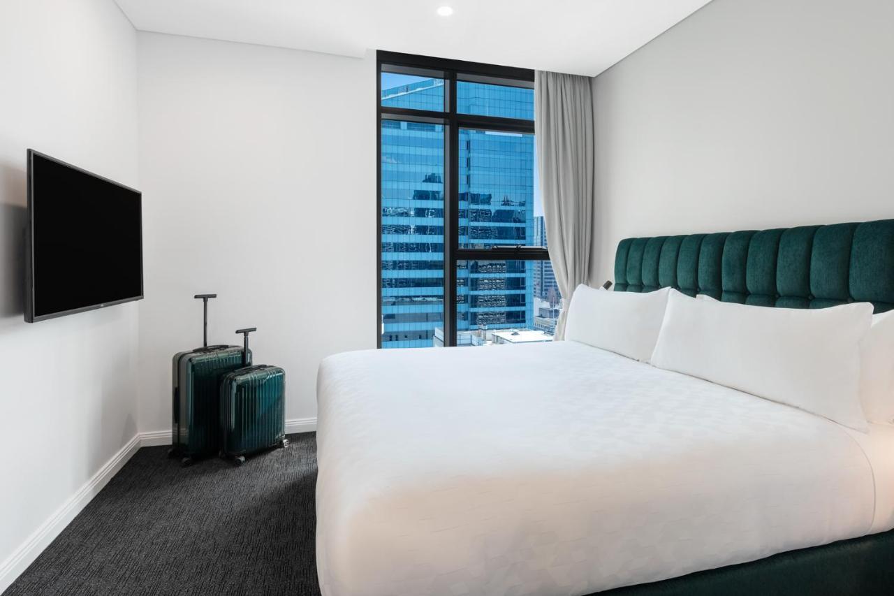 Meriton Suites Sussex Street, Sídney Exterior foto The photo shows a modern hotel room featuring a large bed with white bedding and a green upholstered headboard. There are two green suitcases placed beside the bed. A flat-screen TV is mounted on the wall opposite the bed. The room has a large window