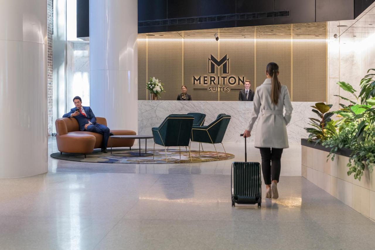Meriton Suites Sussex Street, Sídney Exterior foto The photo depicts a modern hotel lobby. In the foreground, a woman is walking towards the reception desk, pulling a suitcase behind her. She is dressed in a light-colored coat. To her left, there’s a comfortable seating area with a person sitting on 