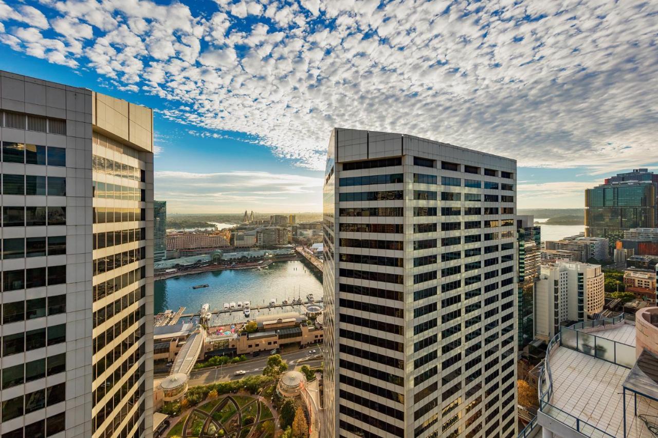 Meriton Suites Sussex Street, Sídney Exterior foto The photo depicts a panoramic view of a modern cityscape featuring tall commercial buildings with glass facades. The shot is taken from a height, showing the intricate architecture of the nearby structures. Below, there is a body of water, which appe