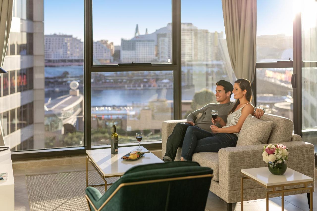Meriton Suites Sussex Street, Sídney Exterior foto The photo shows a cozy indoor setting with a couple sitting closely together on a light-colored sofa. They are holding drinks, likely wine, and appear to be enjoying each other's company while looking out of a large window. The window provides a view
