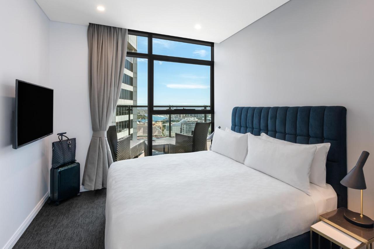 Meriton Suites Sussex Street, Sídney Exterior foto The photo shows a modern hotel room featuring a double bed with white linens and a tufted blue headboard. There is a small bedside table next to the bed. A large window allows natural light to fill the room, offering a view of the city outside, inclu