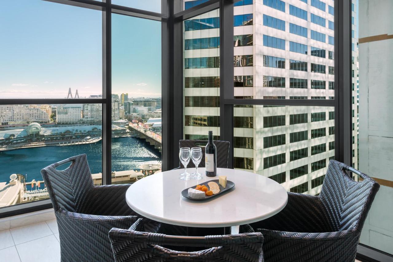 Meriton Suites Sussex Street, Sídney Exterior foto The photo shows a modern dining area with a round table and four woven chairs. On the table, there are two wine glasses, a bottle of wine, and a plate with food items. The large windows provide a view of a cityscape featuring tall buildings and a bod