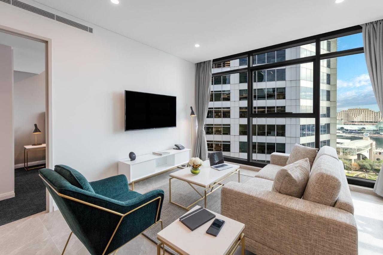 Meriton Suites Sussex Street, Sídney Exterior foto The photo shows a modern living room with large windows that let in natural light, providing a view of a cityscape outside. The interior features a comfortable light-colored sofa and a stylish dark green armchair. A sleek TV is mounted on the wall op