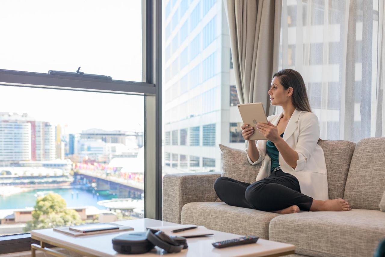 Meriton Suites Sussex Street, Sídney Exterior foto The image portrays a woman sitting cross-legged on a light-colored sofa, engaged with a tablet in her hands. She is dressed in a white blazer and dark pants, appearing relaxed and focused. The setting features large windows that provide a view of a w