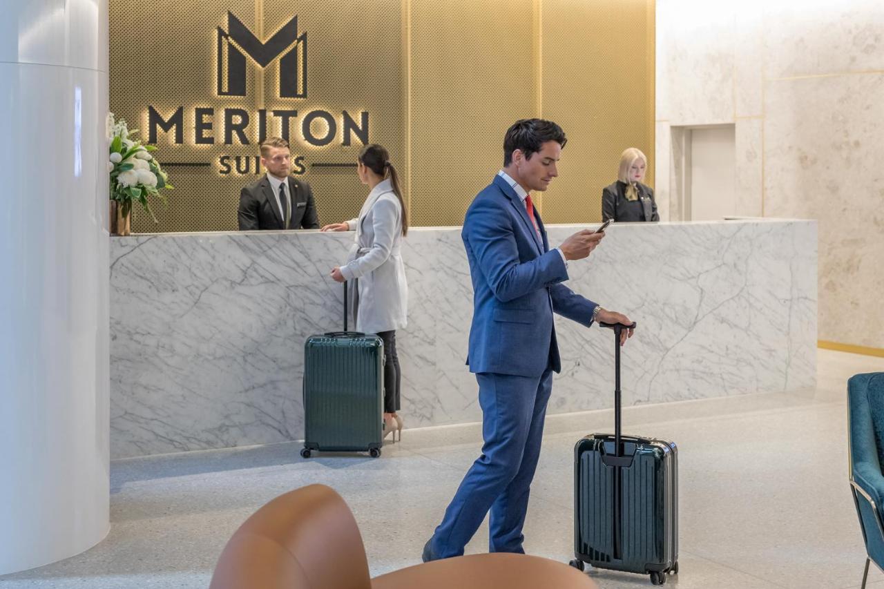 Meriton Suites Sussex Street, Sídney Exterior foto The photo shows the interior of a hotel reception area, specifically at Meriton Suites. In the foreground, a man in a blue suit is standing with a suitcase, looking at his phone. Behind him, there are two staff members at the reception desk, one of w