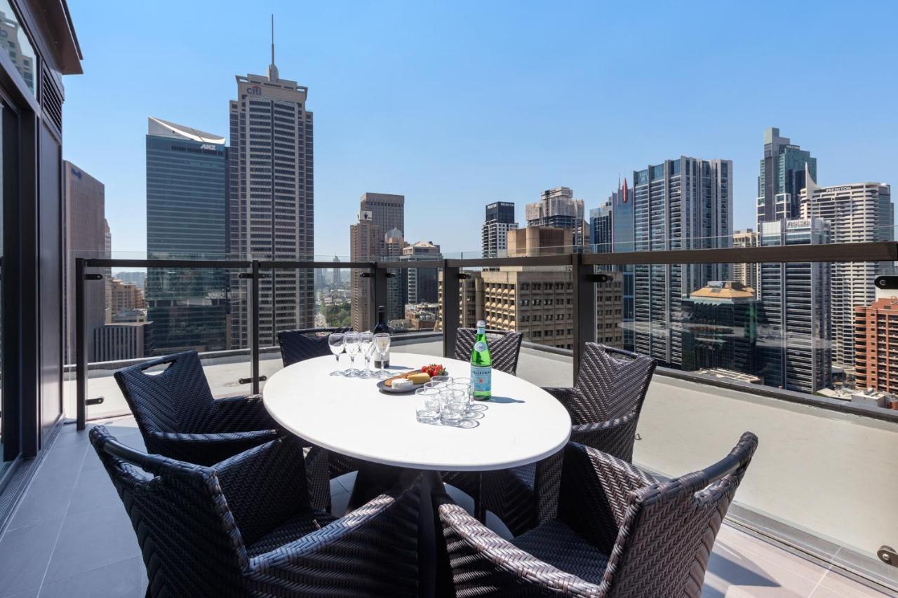 Meriton Suites Sussex Street, Sídney Exterior foto The photo shows a stylish outdoor dining area on a balcony with a round white table surrounded by black wicker chairs. On the table, there are several glasses, a bottle of sparkling water, and a plate of snacks. In the background, there is a panorami