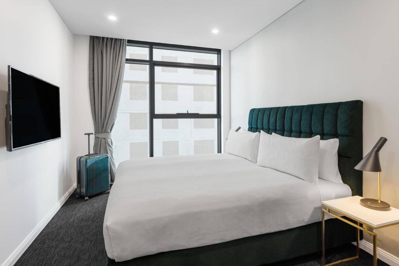 Meriton Suites Sussex Street, Sídney Exterior foto The photo shows a modern hotel room. It features a large bed with a dark green tufted headboard and multiple white pillows. There's a nightstand with a lamp beside the bed. A sleek television is mounted on the wall opposite the bed, and a suitcase is