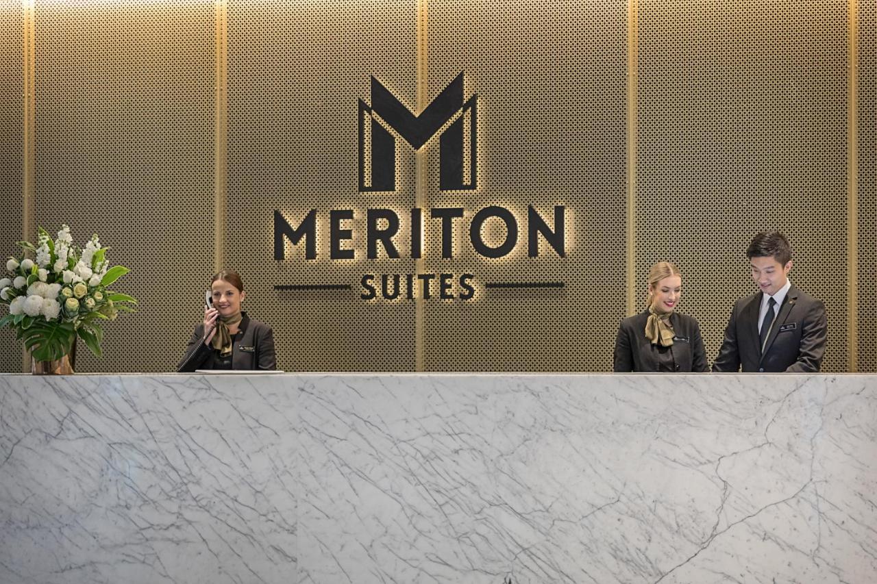 Meriton Suites Sussex Street, Sídney Exterior foto The photo depicts the reception area of a hotel called "Meriton Suites." In the background, there is a large, illuminated sign featuring the hotel’s logo. The reception desk is made of marble and is staffed by two individuals dressed in professional 