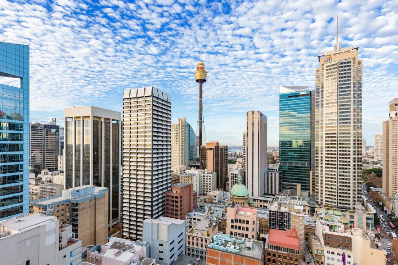 Meriton Suites Sussex Street, Sídney Exterior foto The photo shows a panoramic view of a city skyline dominated by tall skyscrapers. In the center, there is a notable tower with a distinctive golden structure at its top, which is likely a landmark of the city. The buildings have a mix of modern glass