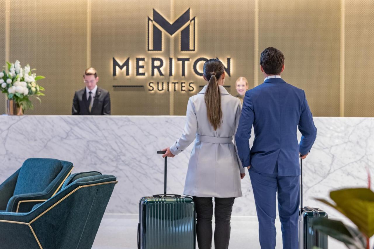 Meriton Suites Sussex Street, Sídney Exterior foto The photo shows a couple standing at a reception area in a hotel named "Meriton Suites." They are both facing the reception desk, where staff members are visible. The man is dressed in a suit, and the woman is wearing a light-colored coat. They each 