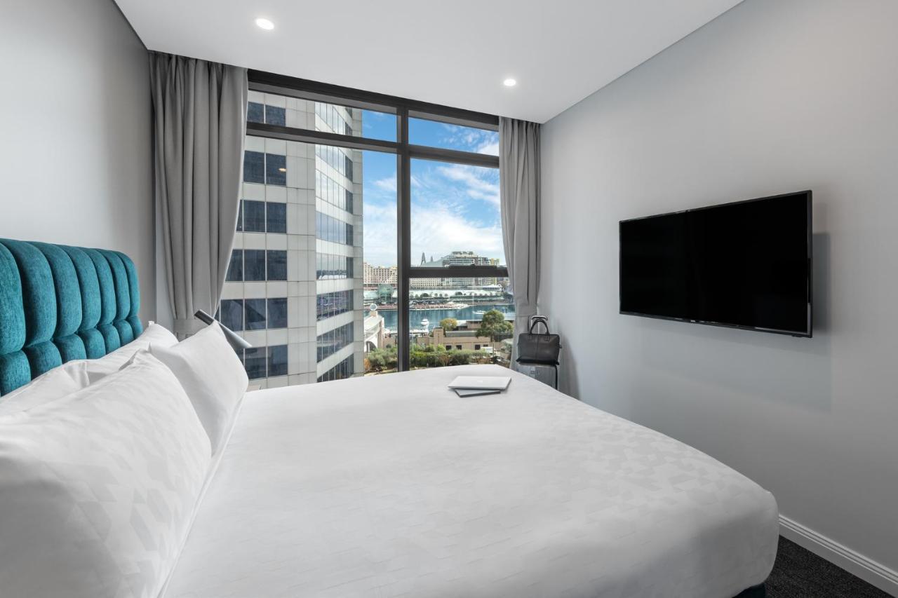 Meriton Suites Sussex Street, Sídney Exterior foto The photo shows a modern hotel room with a large bed featuring a white comforter and pillows. There is a blue headboard that adds a pop of color to the decor. A large window provides a view of the outside, which includes buildings and a glimpse of wa