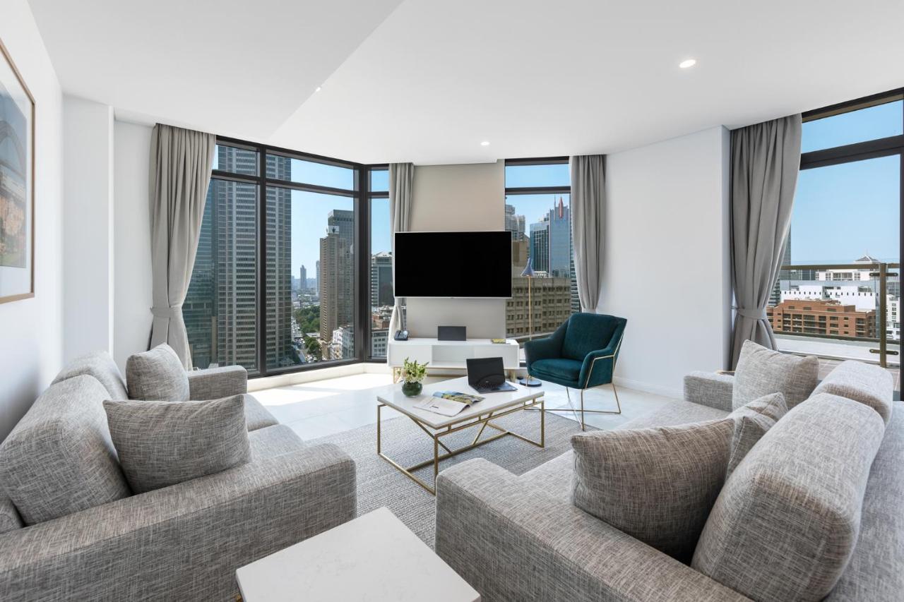 Meriton Suites Sussex Street, Sídney Exterior foto The photo showcases a modern living room with large windows offering a view of a city skyline. The room features a light color palette with gray sofas arranged around a coffee table. A flat-screen TV is mounted on the wall, and there is a green armch