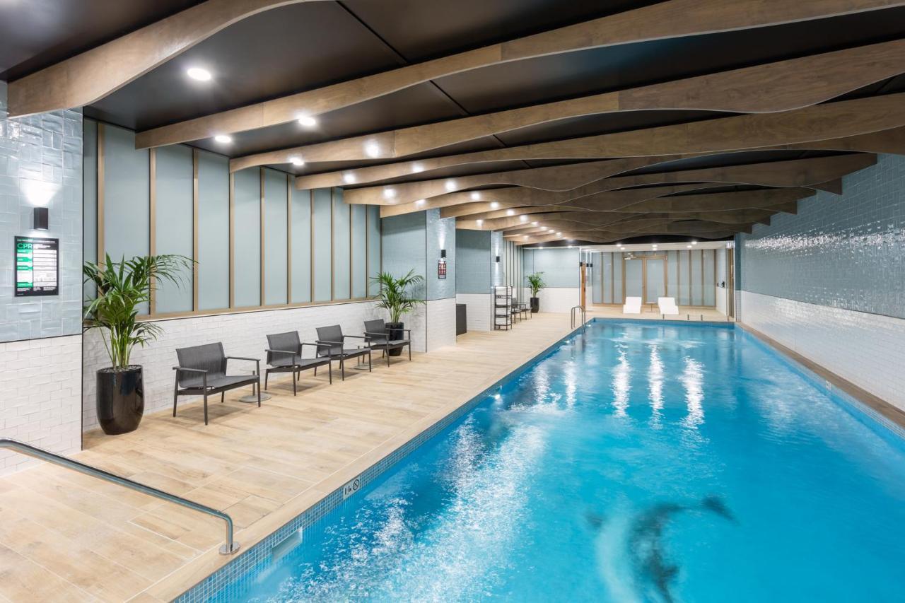 Meriton Suites Sussex Street, Sídney Exterior foto The photo shows an indoor swimming pool area. The pool has clear blue water and is flanked by a wooden deck. There are several lounge chairs arranged along one side of the pool. The ceiling features a modern design with wooden beams and recessed ligh