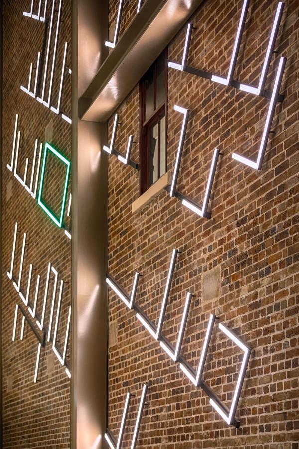 Meriton Suites Sussex Street, Sídney Exterior foto The photo shows a brick wall featuring a design made up of linear light fixtures. These light strips create geometric shapes and patterns, with some sections illuminated in white and one section highlighted in green. The combination of the brick text