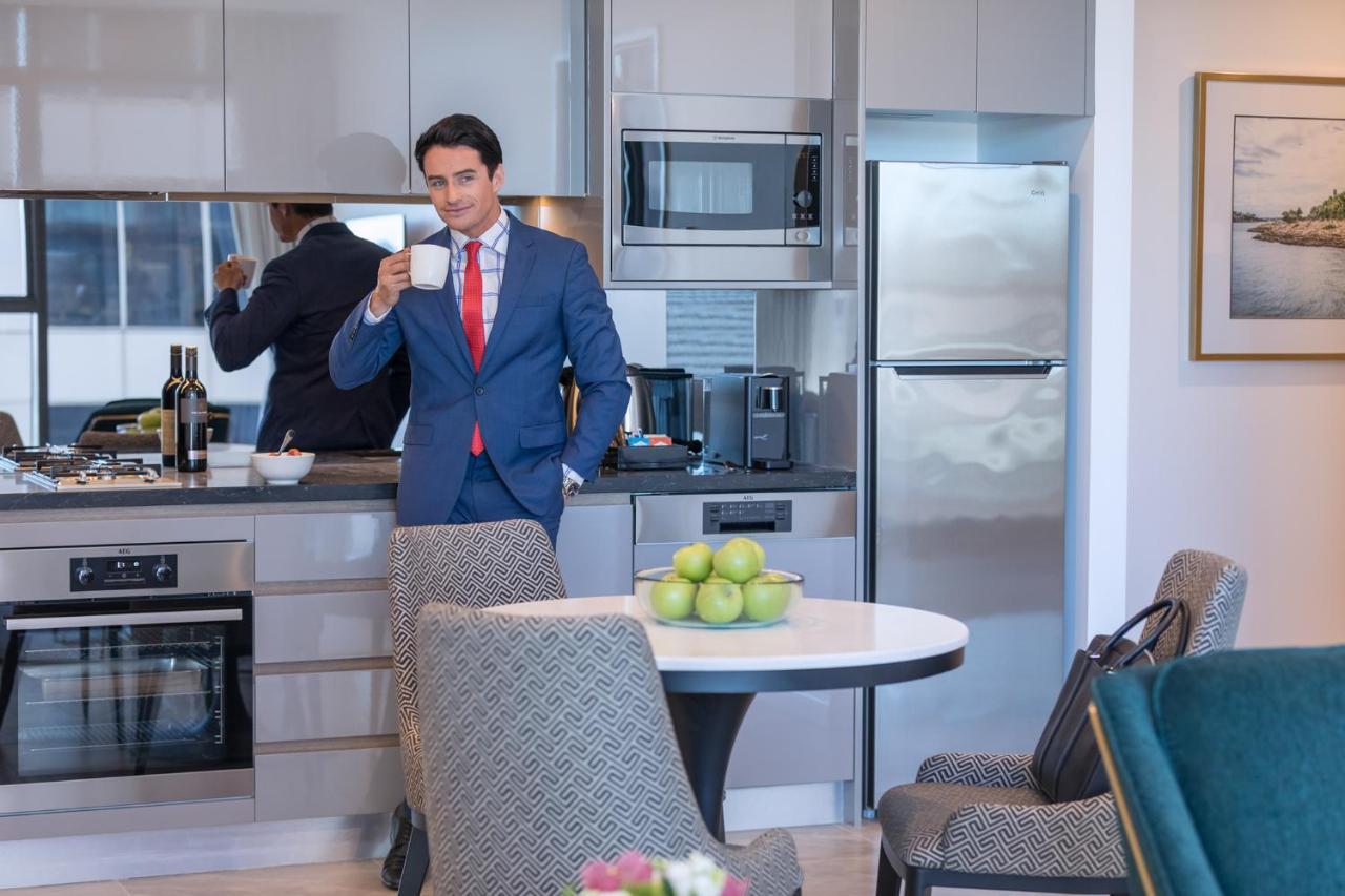 Meriton Suites Sussex Street, Sídney Exterior foto The photo depicts a well-dressed man in a suit standing in a modern kitchen. He is holding a cup, possibly of coffee, and appears to be looking towards the camera. The kitchen features contemporary appliances, including a microwave and refrigerator. 