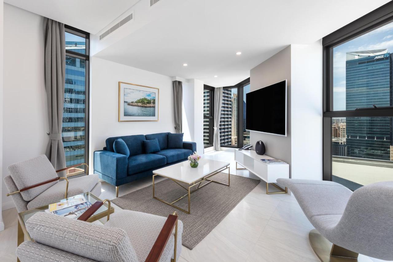 Meriton Suites Sussex Street, Sídney Exterior foto The photo shows a modern living room featuring large windows that allow plenty of natural light. The room is elegantly designed with a blue sofa, a light-colored coffee table, and two armchairs. There’s a flat-screen TV mounted on the wall, and a pie